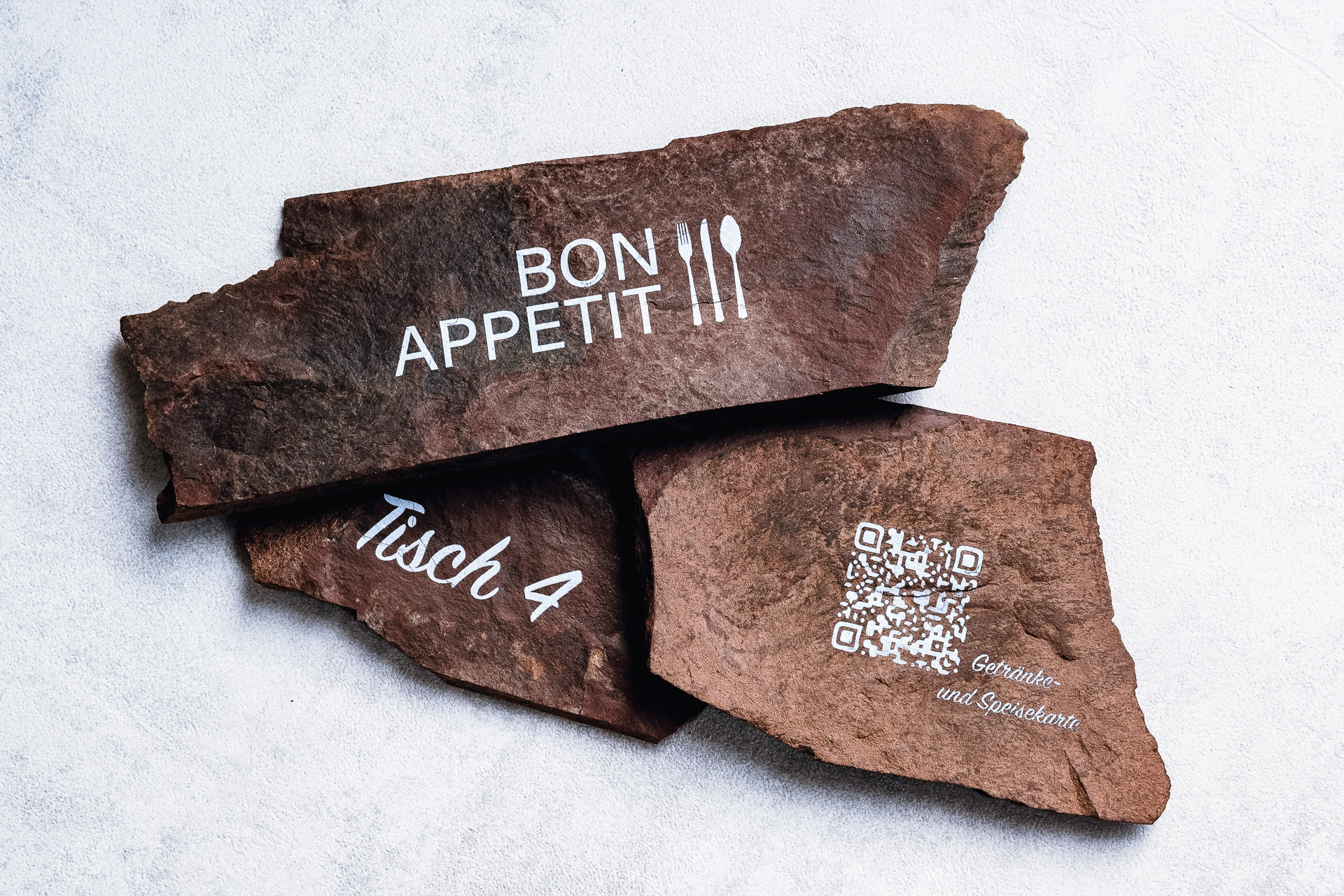 Bedruckte Schieferplatten für die Gastronomie, beschriftet mit "Bon Apetit", "Tisch 4" und einem QR-Code zum Scannen